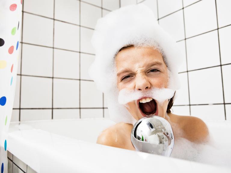 Ein Junge sitzt in der Badewanne mit Schaum im Gesicht und singt in einen Duschkopf.