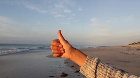 Ein ausgestreckter Arm vor einem Strandpanorama, den Daumen nach oben gereckt. 