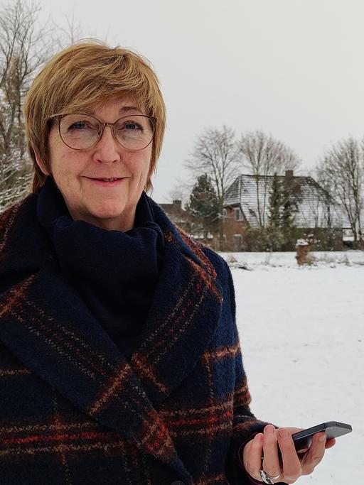 Elke Vespermann steht mit ihrem Smartphone in der Hand in einer verschneiten Landschaft. Sie trägt einen Mantel, Brille und schaut freundlich in die Kamera