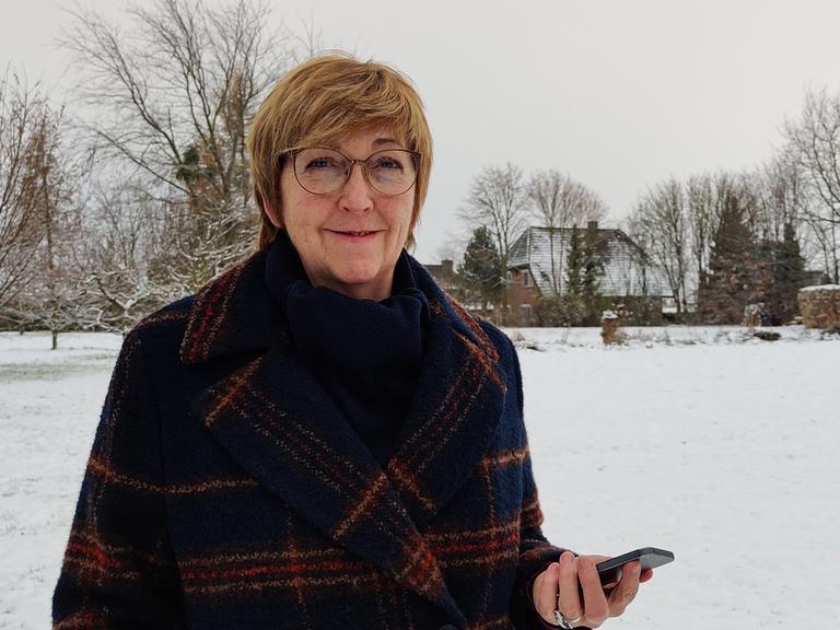 Elke Vespermann steht mit ihrem Smartphone in der Hand in einer verschneiten Landschaft. Sie trägt einen Mantel, Brille und schaut freundlich in die Kamera