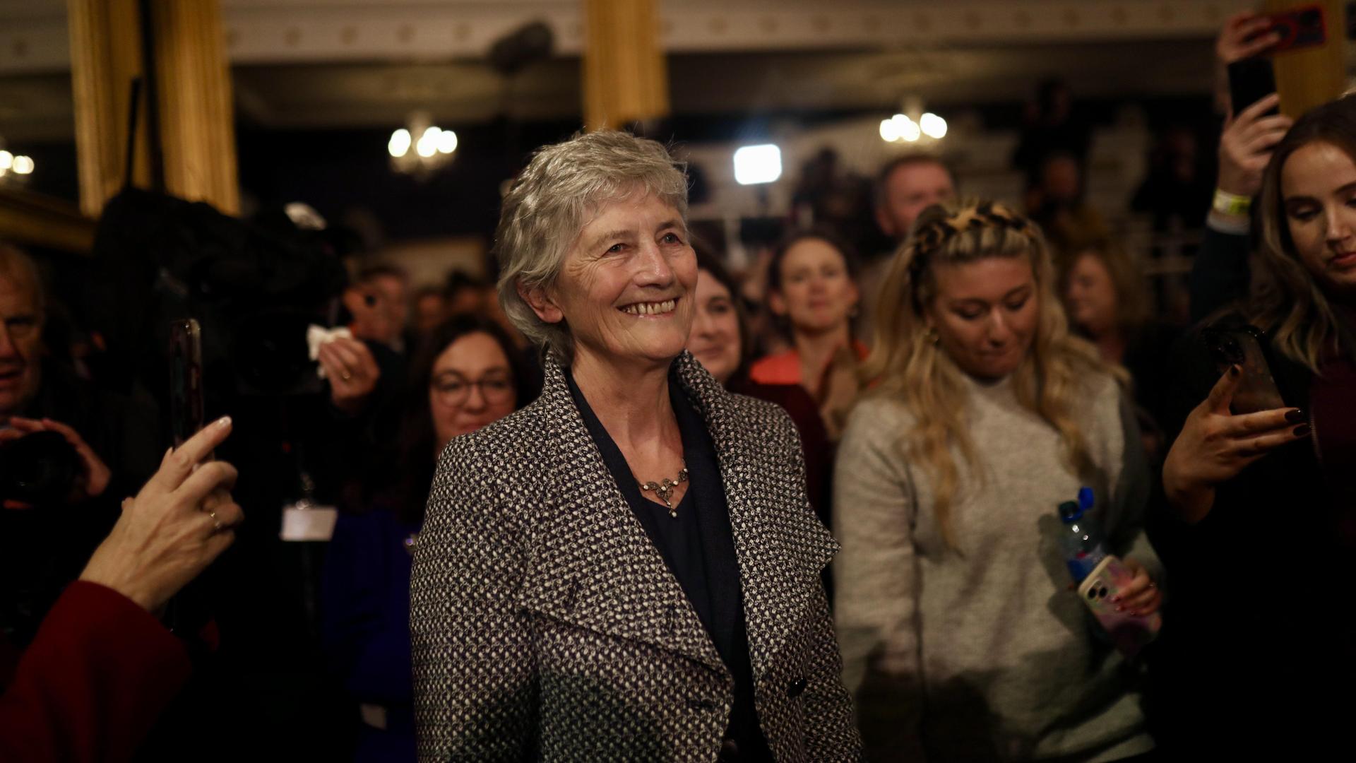 Catherine Connolly steht in einer Gruppe von Menschen. Sie trägt einen grauen Mantel. Catherine Connolly steht in einer Gruppe von Menschen. Sie trägt einen grauen Mantel.