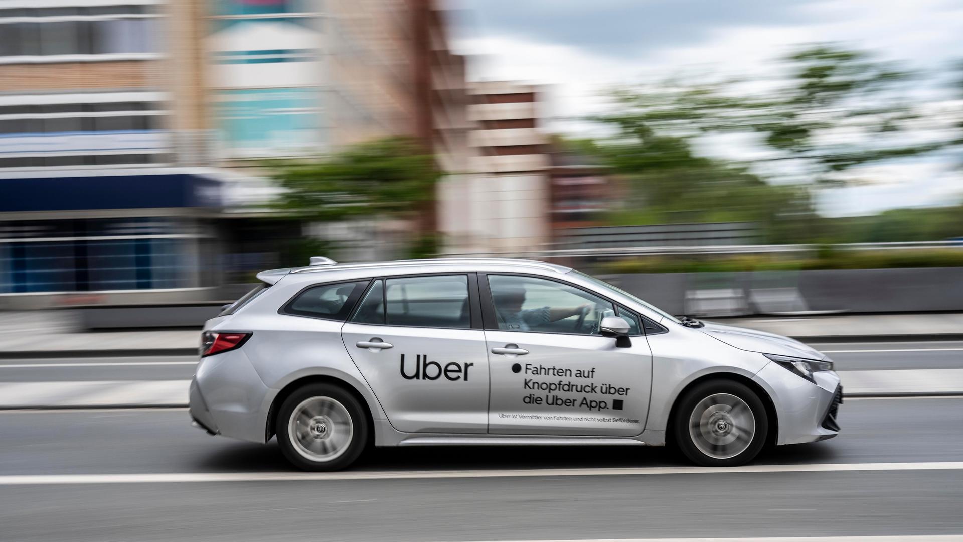 Blick von der Seite auf das silbergraue Auto mit der Uber-Aufschrift an der Seite, das eine Straße entlangfährt. Der Hintergrund zeigt eine Häuserzeile und ist wegen der Geschwindigkeit unscharf. Blick von der Seite auf das silbergraue Auto mit der Uber-Aufschrift an der Seite, das eine Straße entlangfährt. Der Hintergrund zeigt eine Häuserzeile und ist wegen der Geschwindigkeit unscharf.