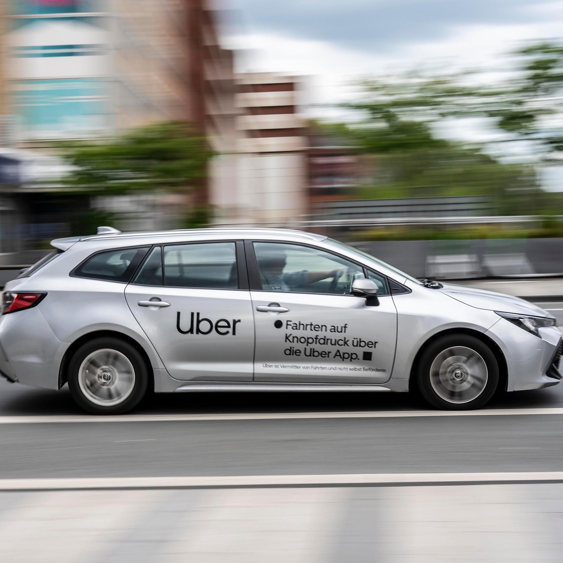 Blick von der Seite auf das silbergraue Auto mit der Uber-Aufschrift an der Seite, das eine Straße entlangfährt. Der Hintergrund zeigt eine Häuserzeile und ist wegen der Geschwindigkeit unscharf. 
