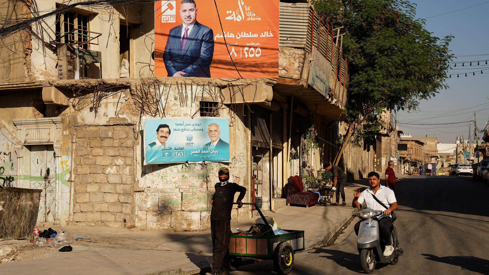 Ein Junge mit einem Karren und ein Mopedfahrer vor Wahlplakaten der politischen Kandidaten Khaled Sultan Hashem und Rayan Ahmed Al-Omar in der Stadt Mossul Ein Junge mit einem Karren und ein Mopedfahrer vor Wahlplakaten der politischen Kandidaten Khaled Sultan Hashem und Rayan Ahmed Al-Omar in der Stadt Mossul
