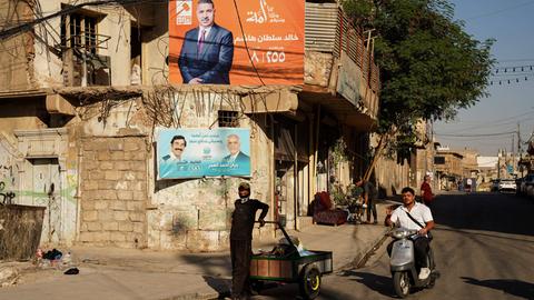 Ein Junge mit einem Karren und ein Mopedfahrer vor Wahlplakaten  der politischen Kandidaten Khaled Sultan Hashem und Rayan Ahmed Al-Omar in der Stadt Mossul