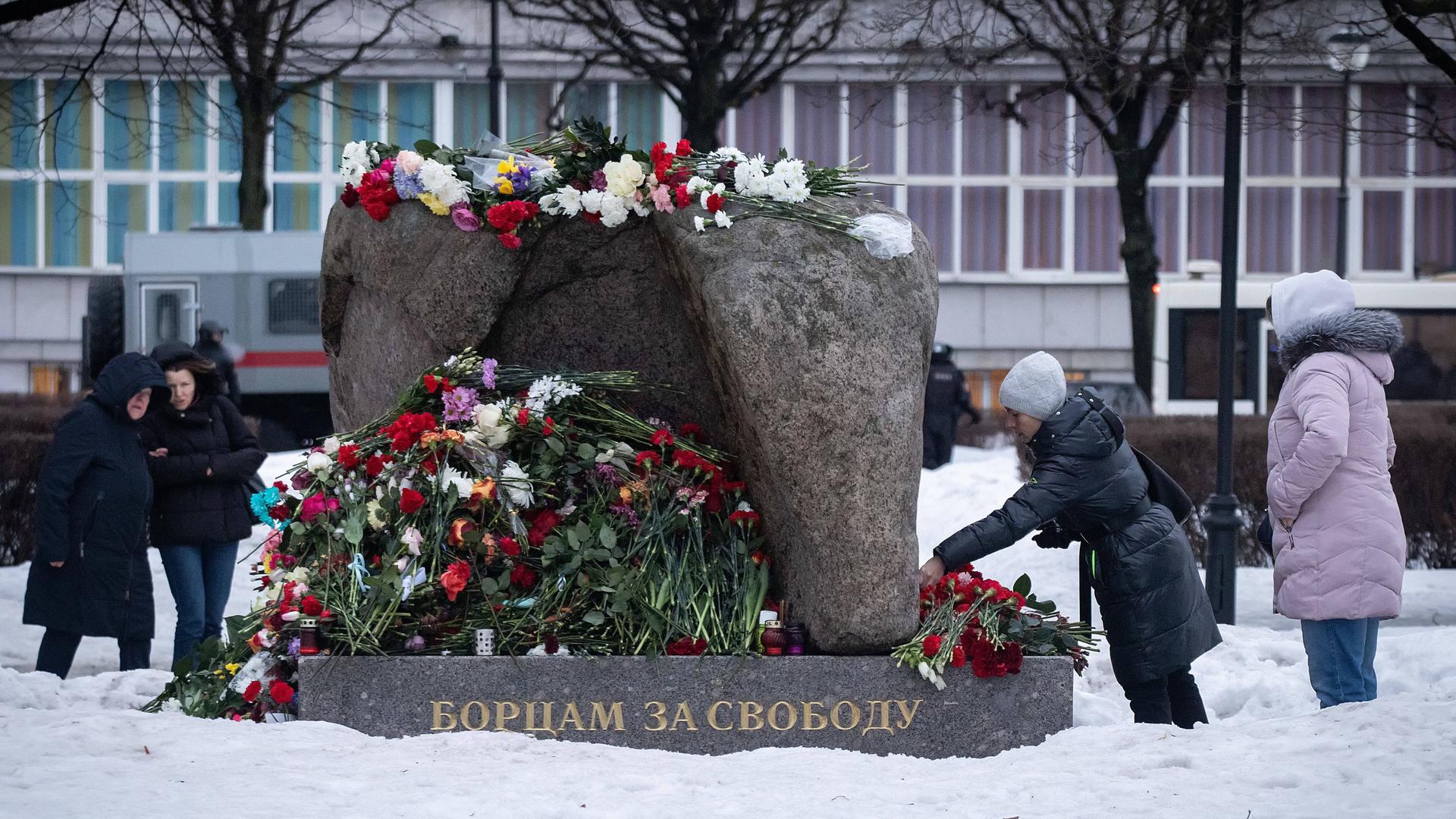 An einem Denkmal in der russischen Stadt St. Petersburg liegen viele Blumen. Eine Frau legt weitere Blumen ab. Rings um das Denkmal stehen einige Menschen.