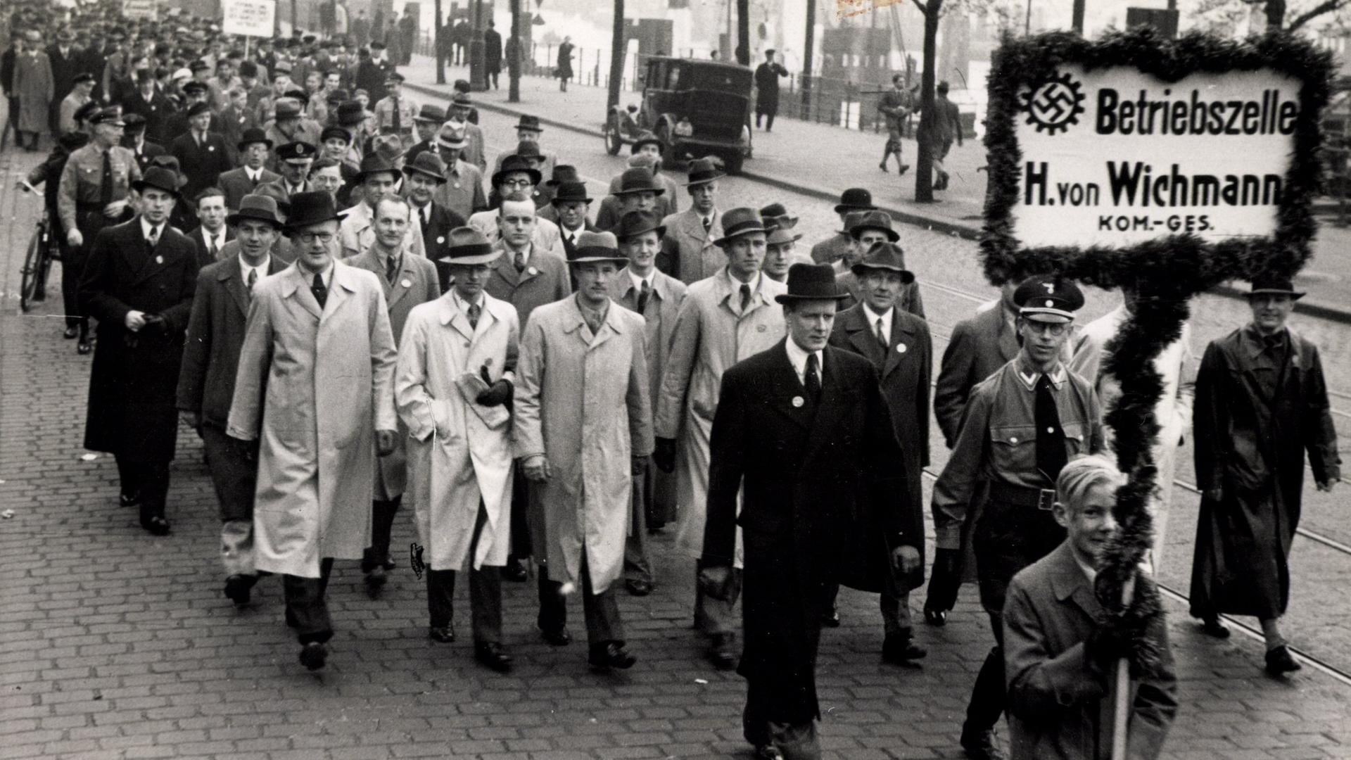 Eine Gruppe von Männern in Mäntel läuft zusammen eine Straße entlang. Der Gruppe voran gestellt läuft ein Junge, der ein Schild mit Hakenkreuz trägt auf dem "Betriebszelle H. von Wichmann" zu lesen ist.
