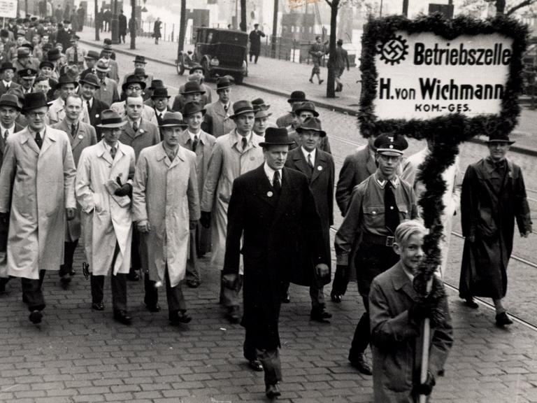 Eine Gruppe von Männern in Mäntel läuft zusammen eine Straße entlang. Der Gruppe voran gestellt läuft ein Junge, der ein Schild mit Hakenkreuz trägt auf dem "Betriebszelle H. von Wichmann" zu lesen ist.