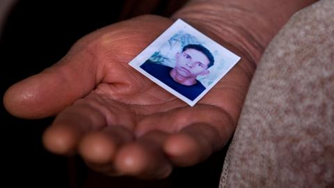 Nahaufnahme einer Frauenhand, die das Passfoto eines jungen Mannes in die Kamera hält. Er hat kurzes lockiges Haar und schaut ernst.