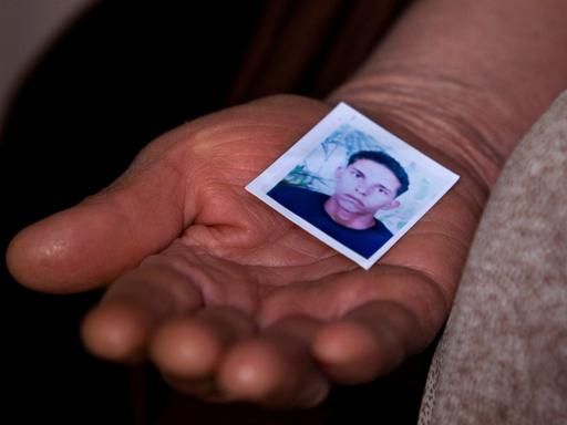 Nahaufnahme einer Frauenhand, die das Passfoto eines jungen Mannes in die Kamera hält. Er hat kurzes lockiges Haar und schaut ernst.