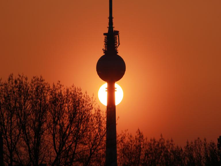 Der Berliner Fernsehturm vor der untergehenden Sonne, die tiefrot am Himmel steht.