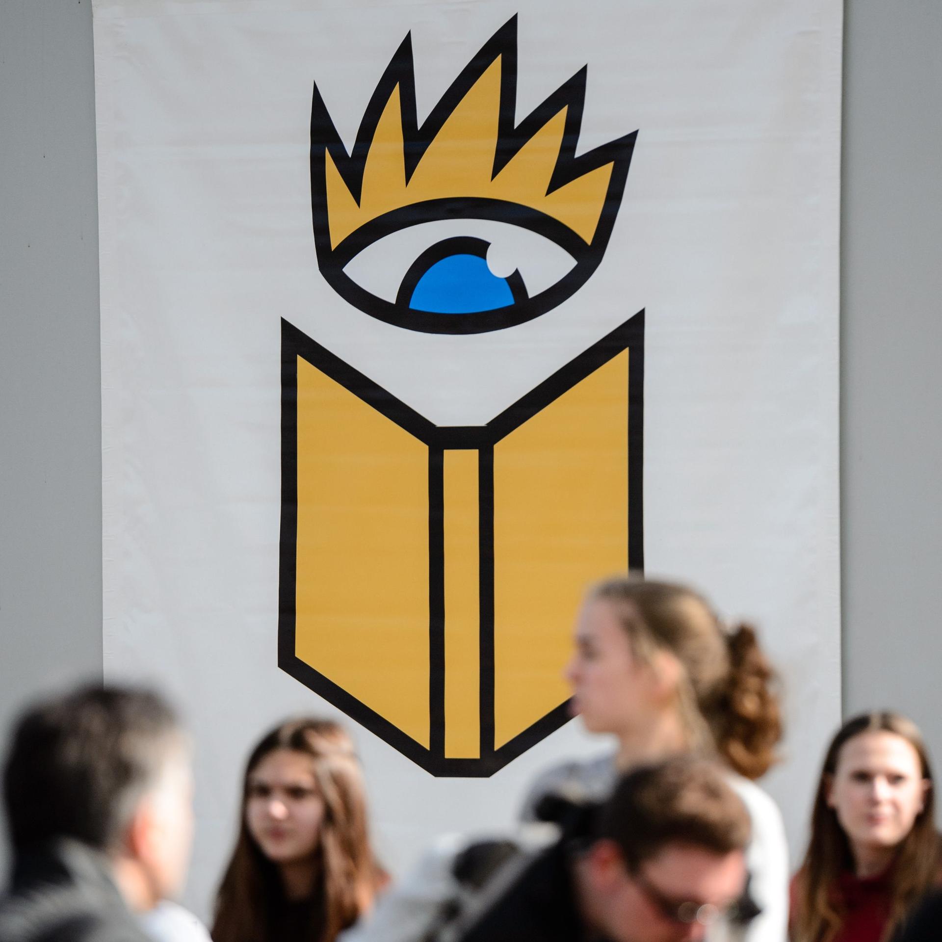 Besucher der Leipziger Buchmesse gehen unter dem Logo der Messe hin und her.