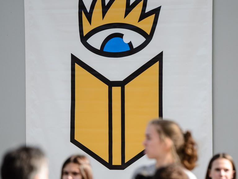 Besucher der Leipziger Buchmesse gehen unter dem Logo der Messe hin und her.