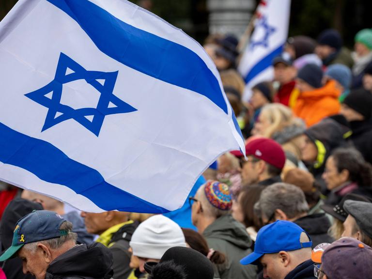 Menschen stehen nebeinander bei einer Kundgebung, einge tragen israelische Flaggen.