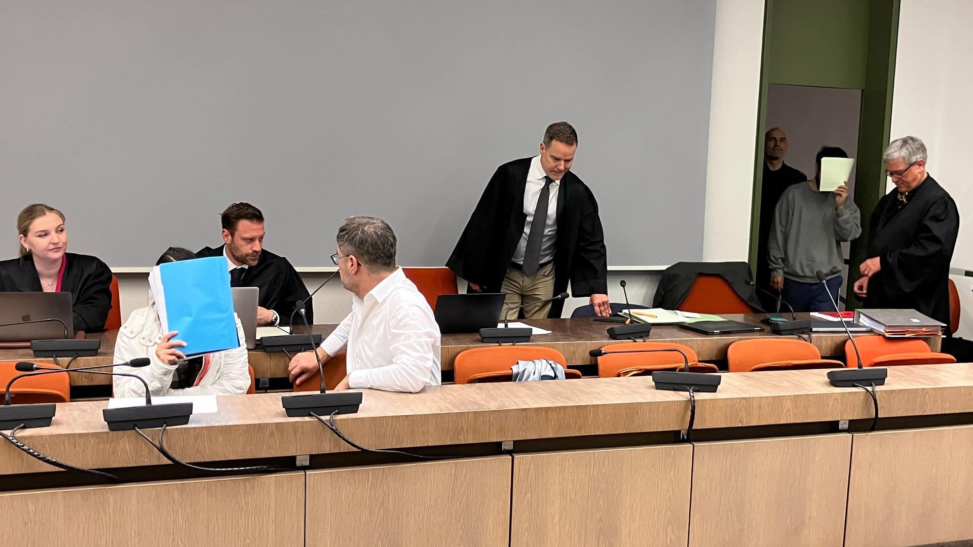 Die wegen Versklavung, Vergewaltigung und Verbrechen gegen die Menschlichkeit angeklagte irakische Frau (2.v.l mit Mappe vor dem Gesicht) sitzt im Saal des Oberlandesgerichts, während ihr mitangeklagter Mann (hinten, 2.v.r) ebenfalls in den Saal kommt. 