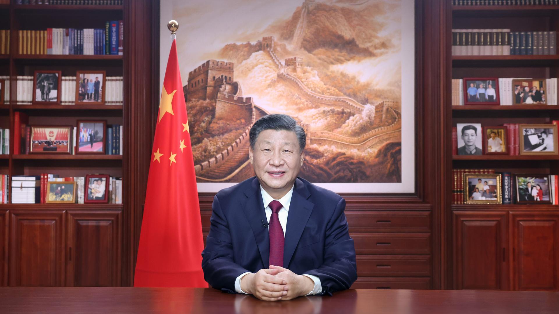 Chinas Staatschef Xi Jinping sitzt an einem Schreibtisch. Neben ihm sieht man die chinesische Flagge, im Hintergrund ist ein großes Bild mit der chinesischen Mauer.