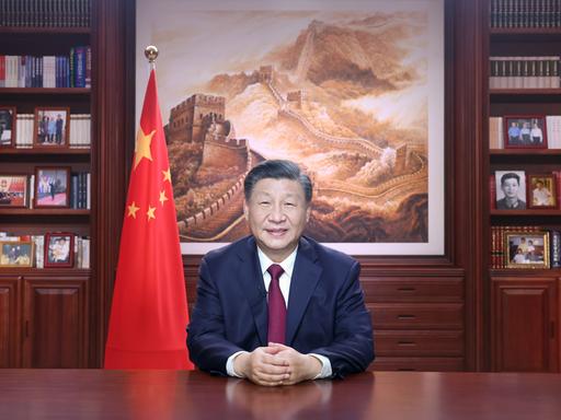 Chinas Staatschef Xi Jinping sitzt an einem Schreibtisch. Neben ihm sieht man die chinesische Flagge, im Hintergrund ist ein großes Bild mit der chinesischen Mauer.