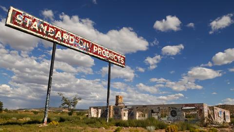 Vor blauem Himmel ist eine verlassene Tankstelle zu sehen. Davor steht ein rostiges Schild "Standard Oil Products".
