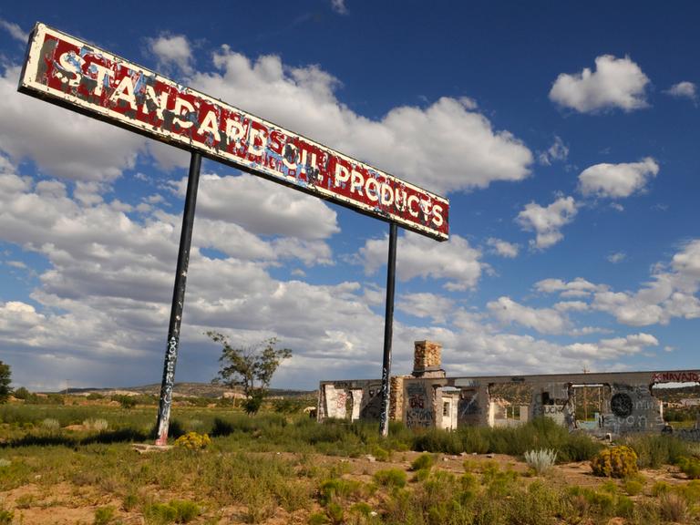 Vor blauem Himmel ist eine verlassene Tankstelle zu sehen. Davor steht ein rostiges Schild "Standard Oil Products".