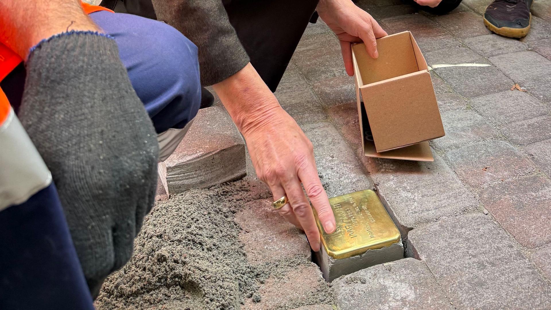 Zu sehen sind die Hände der CDU-Politikerin, die den golden glänzenden Pflasterstein auf einem Bürgersteig in ein vorbereitetes Loch legt. 