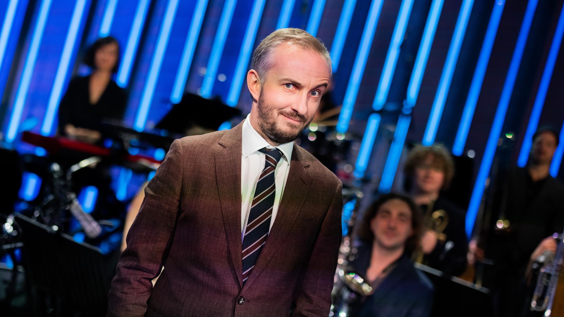 Jan Böhmermann steht im Studio des ZDF Magazin Royal vor seinem Orchester und macht eine ironische Grimasse mit hochgezogener Augenbraue.