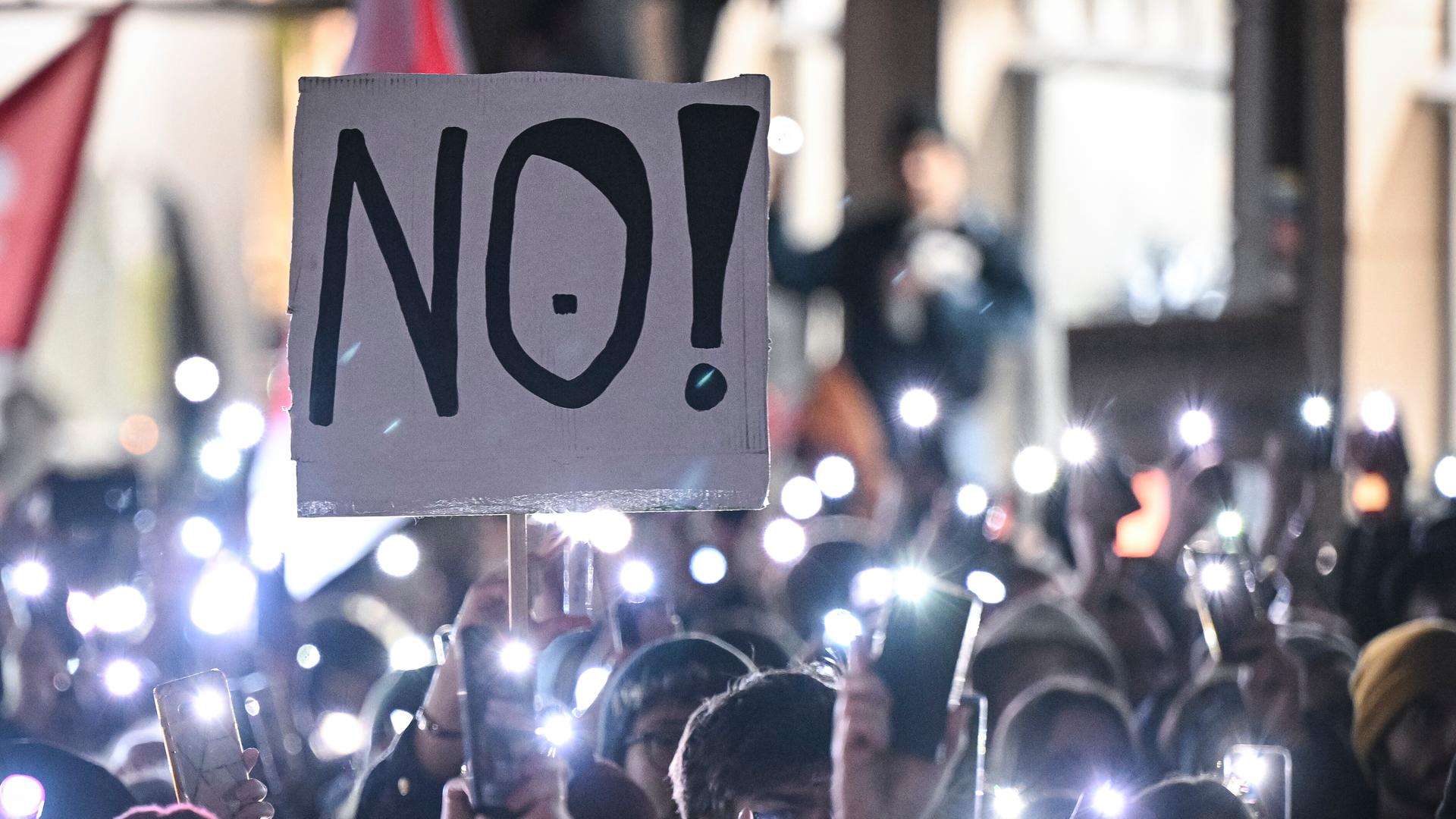 Auf einer Demonstration in Tübingen stehen Menschen eng aneinander und halten ihre leuchtenden Handy-Taschenlampen in die Luft. Eine Person hält ein Schild mit der Aufschrift "NO!" in die Luft.