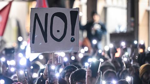 Auf einer Demonstration in Tübingen stehen Menschen eng aneinander und halten ihre leuchtenden Handy-Taschenlampen in die Luft. Eine Person hält ein Schild mit der Aufschrift "NO!" in die Luft.
