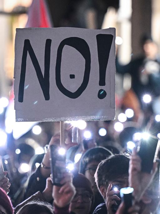 Auf einer Demonstration in Tübingen stehen Menschen eng aneinander und halten ihre leuchtenden Handy-Taschenlampen in die Luft. Eine Person hält ein Schild mit der Aufschrift "NO!" in die Luft.
