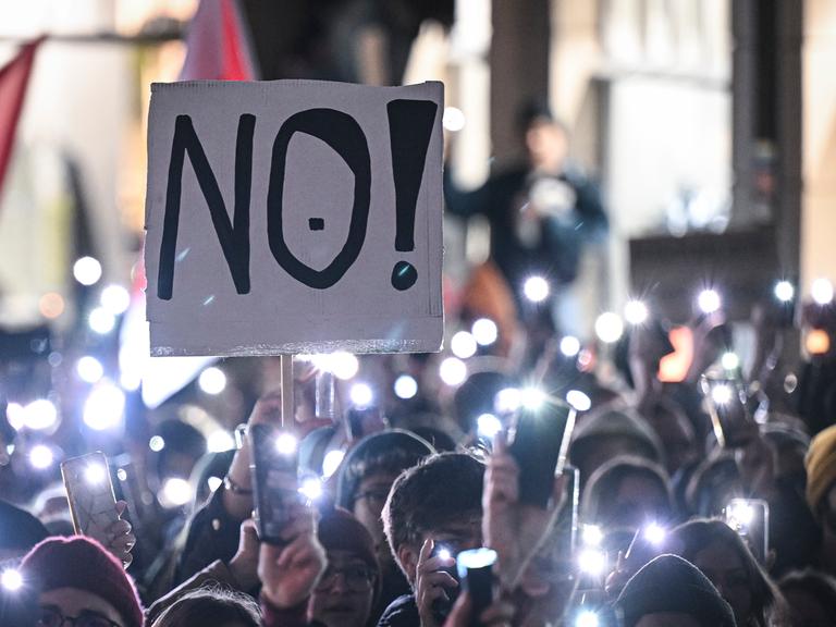 Auf einer Demonstration in Tübingen stehen Menschen eng aneinander und halten ihre leuchtenden Handy-Taschenlampen in die Luft. Eine Person hält ein Schild mit der Aufschrift "NO!" in die Luft.