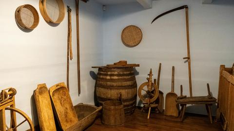 Blick in einen kellerartigen Raum in dem historische Landwirtschaftsgeräte stehen und an den Wänden hängen wie ein Spinnrad und Sensen.