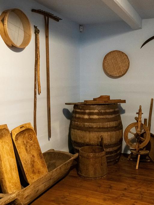 Blick in einen kellerartigen Raum in dem historische Landwirtschaftsgeräte stehen und an den Wänden hängen wie ein Spinnrad und Sensen.
