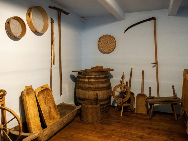 Blick in einen kellerartigen Raum in dem historische Landwirtschaftsgeräte stehen und an den Wänden hängen wie ein Spinnrad und Sensen.