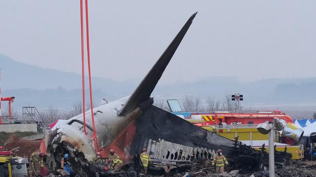 Helfer arbeiten vor dem kaputten  Flugzeug. Das Flugzeug ist in viele Teile zerbrochen und ausgebrannt. Ein Flügel ragt in den Himmel.