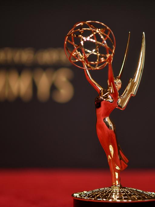 Vor einem verschwommenen "Emmy"-Schriftzug steht die Trophäe der Emmy Awards, eine Frauenstatue mit Flügeln, die einen geflochtenen Draht-Ball in die Höhe hält.