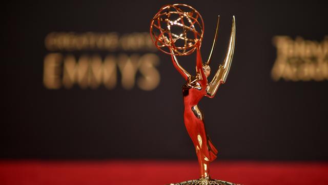 Vor einem verschwommenen "Emmy"-Schriftzug steht die goldene Trophäe der Emmy Awards:  eine Frauenstatue mit Flügeln, die einen geflochtenen Draht-Ball in die Höhe hält.