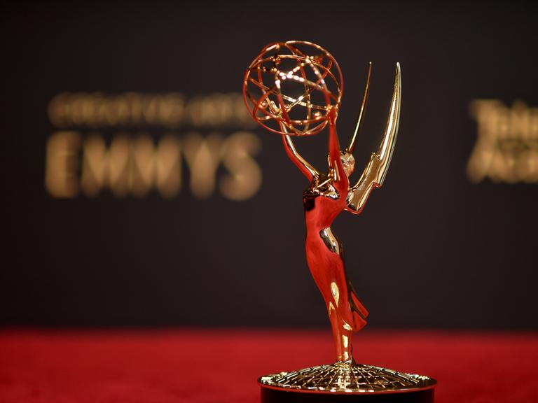 Vor einem verschwommenen "Emmy"-Schriftzug steht die Trophäe der Emmy Awards, eine Frauenstatue mit Flügeln, die einen geflochtenen Draht-Ball in die Höhe hält.