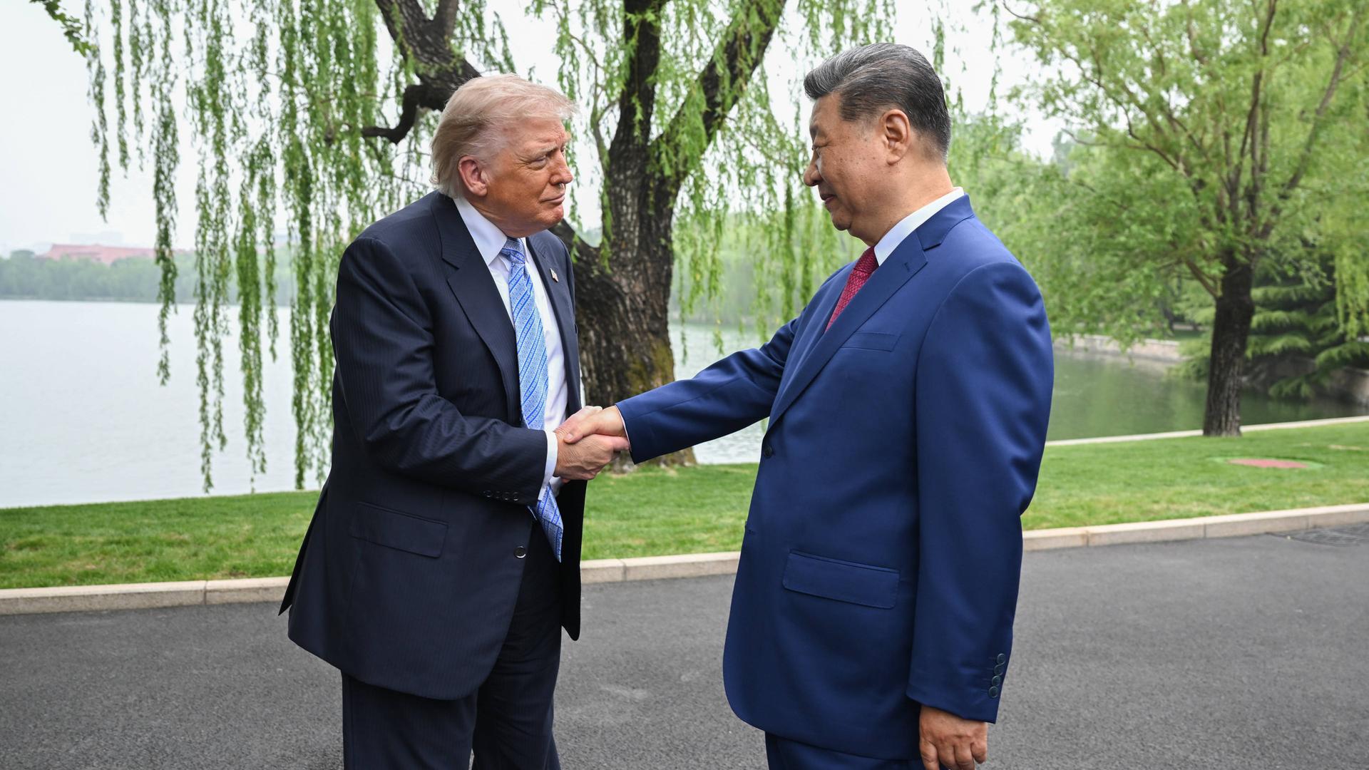 Donald Trump und Xi Jinping schütteln sich die Hände. Beide tragen blaue Anzüge. Im Hintergrund ein Baum und ein Teich.