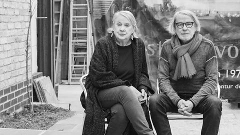 Ute und Werner Mahler sitzen auf zwei Stühlen in einem Innenhof. Sie trägt eine lange schwarze Strickjacke und große Ohrringe. Er trägt einen hellen Schal und eine schwarz umrandete Brille. 