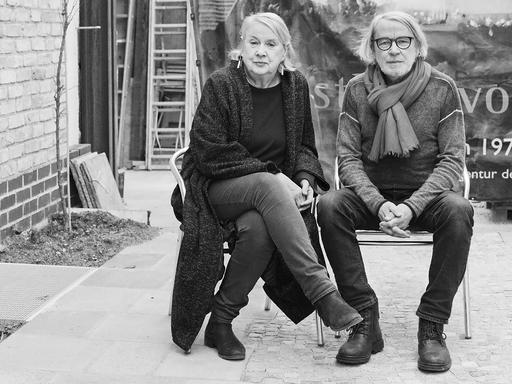 Ute und Werner Mahler sitzen auf zwei Stühlen in einem Innenhof. Sie trägt eine lange schwarze Strickjacke und große Ohrringe. Er trägt einen hellen Schal und eine schwarz umrandete Brille. 