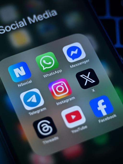 Auf einem Smartphone-Display sind verschiedene Social-Media-Apps zu sehen, darunter WhatsApp, Telegram, Instagram, X und Facebook.