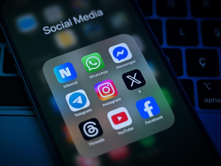 Auf einem Smartphone-Display sind verschiedene Social-Media-Apps zu sehen, darunter WhatsApp, Telegram, Instagram, X und Facebook.