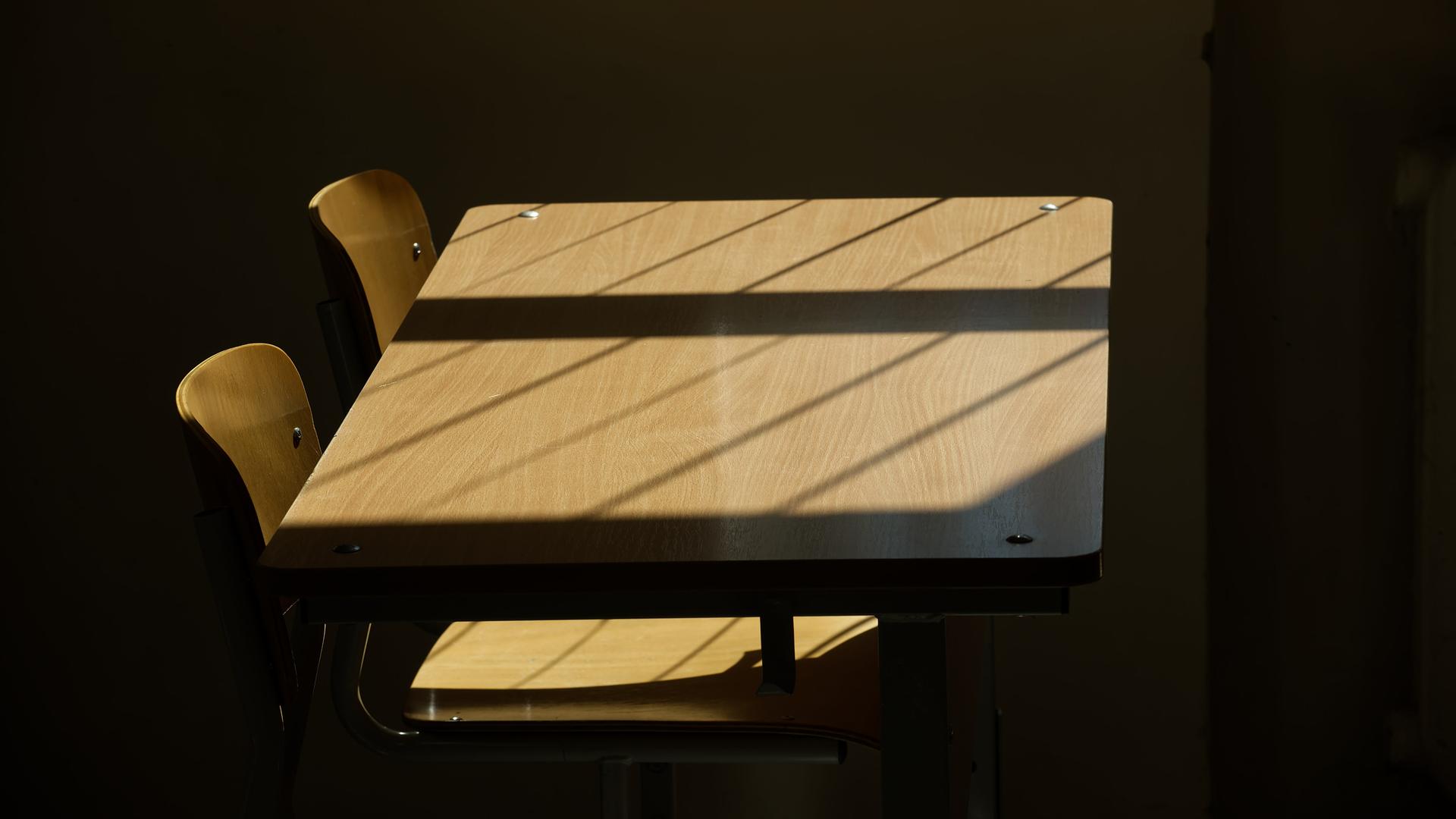 Leere Tische und Stühle in einem Klassenzimmer, durch die Sonne werden Schatten auf das Holz geworfen.