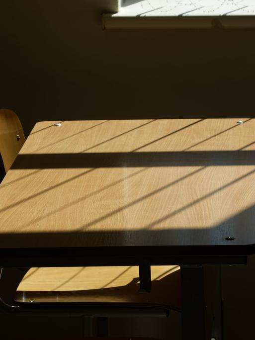 Leere Tische und Stühle in einem Klassenzimmer, durch die Sonne werden Schatten auf das Holz geworfen.