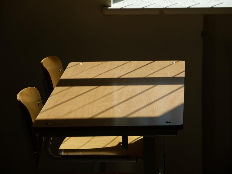 Leere Tische und Stühle in einem Klassenzimmer, durch die Sonne werden Schatten auf das Holz geworfen.