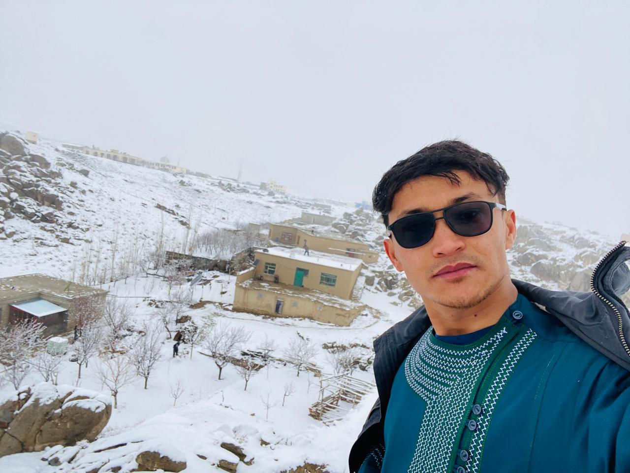 Khadem Ali Azad vor seinem schneebedeckten Heimatdorf in der afghanischen Provinz Dschaghuri. Im Hintergrund sind flache Gebäude und Hügel zu sehen.