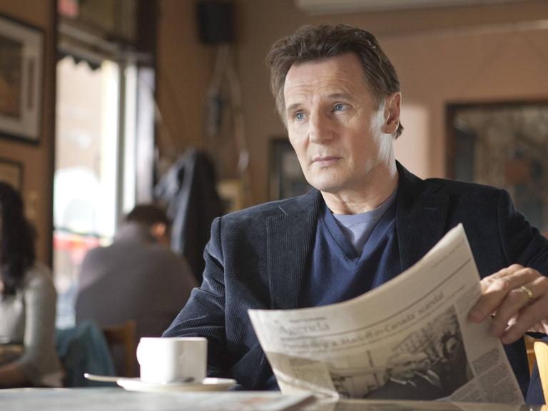 Filmausschnitt von Liam Neeson in "Chloe" wie er in einem Café sitzt und Zeitung liest.