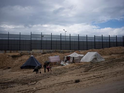 Palästinenser, die durch die israelische Luft- und Bodenoffensive auf den Gazastreifen vertrieben wurden, gehen durch ein behelfsmäßiges Zeltlager in Rafah an der Grenze zu Ägypten. Im Hintergrund ein hoher Grenzzaun.