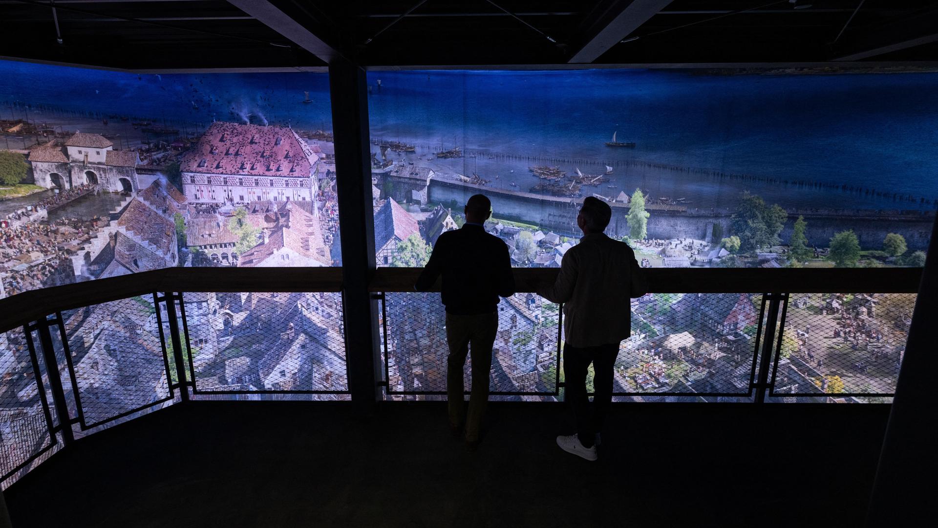 Eine Rundum-Projektion der Stadt Konstanz, wie sie im Mittelalter wohl ausgesehen haben könnte. Man sieht vor der Projekt die Silhouette von zwei Männern, die auf einer Plattform stehen und sich das Bild anschauen. 