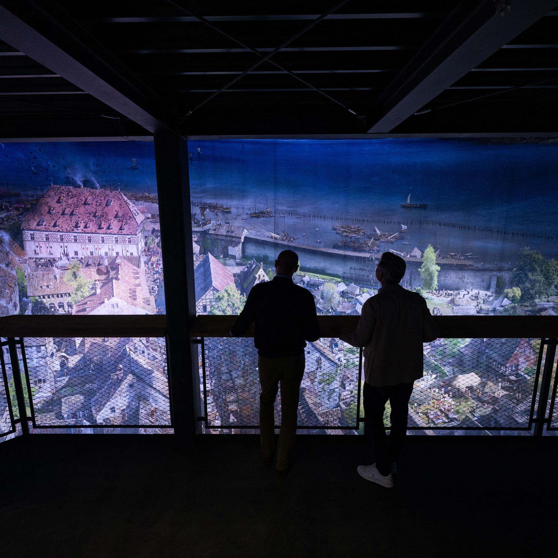 Eine Rundum-Projektion der Stadt Konstanz, wie sie im Mittelalter wohl ausgesehen haben könnte. Man sieht vor der Projekt die Silhouette von zwei Männern, die auf einer Plattform stehen und sich das Bild anschauen. 