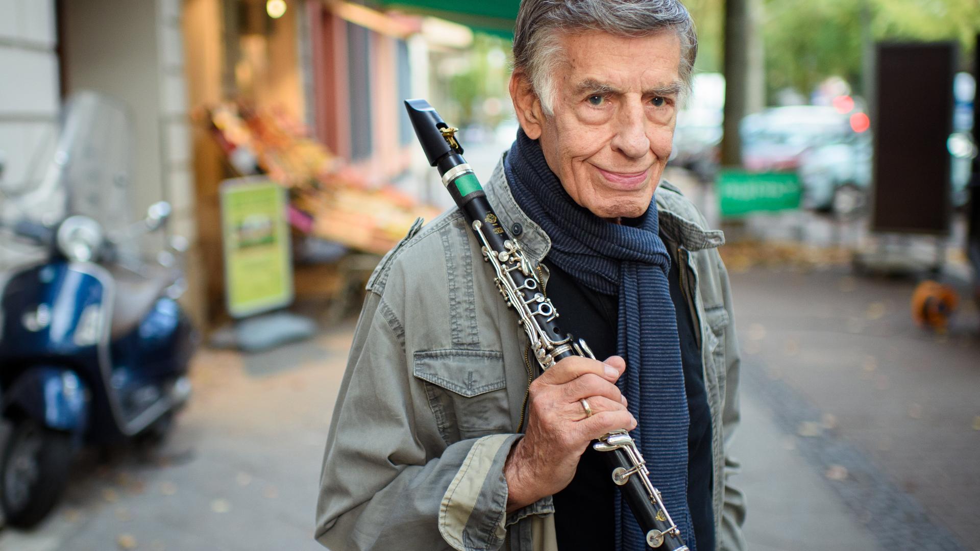 Rolf Kühn, Jazzmusiker und Komponist, steht im Berliner Bezirk Charlottenburg auf einem Gehweg und hält seine Klarinette in der Hand. Er hat graue, zur Seite gekämmte Haare und trägt einen blauen Schal um den Hals.